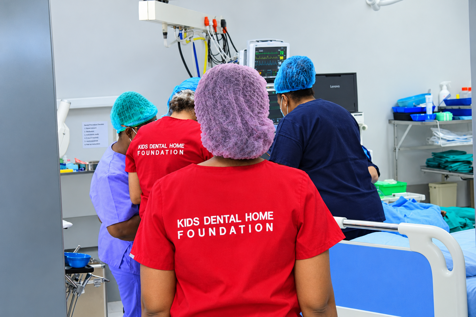 Foundation volunteers in red Kids Dental Home Foundation scrubs, seen from behind, working with a surgical team in an operating theatre